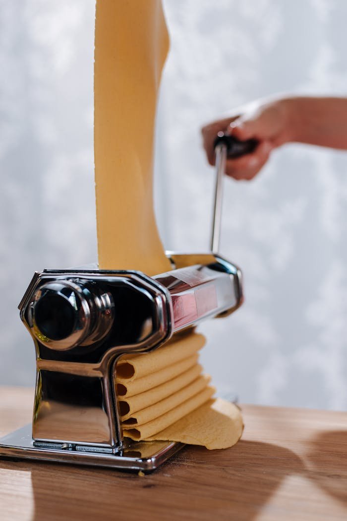 Close-up of a hand using a pasta maker to roll fresh dough on a wooden table.
