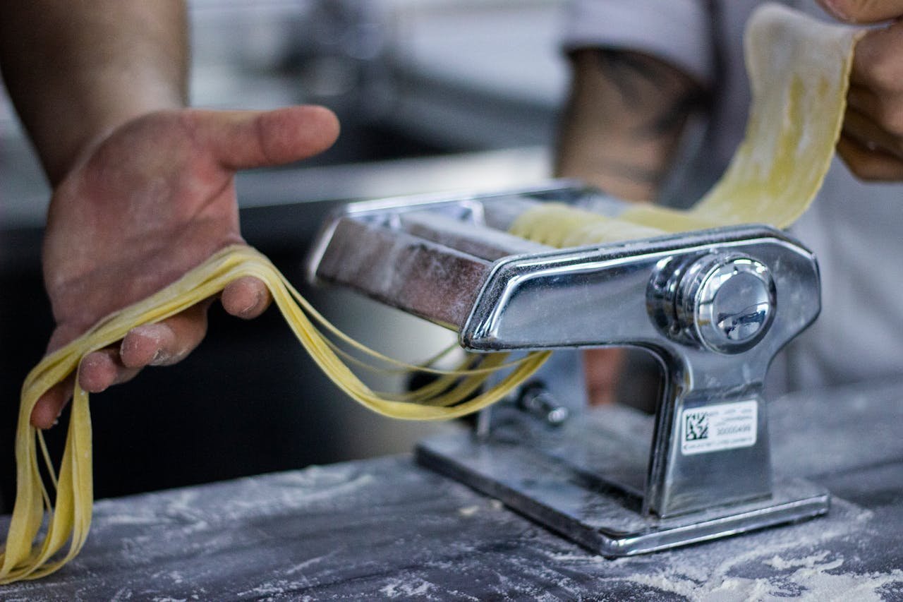 Close-up of fresh pasta being prepared with a manual pasta machine by hands in a kitchen.