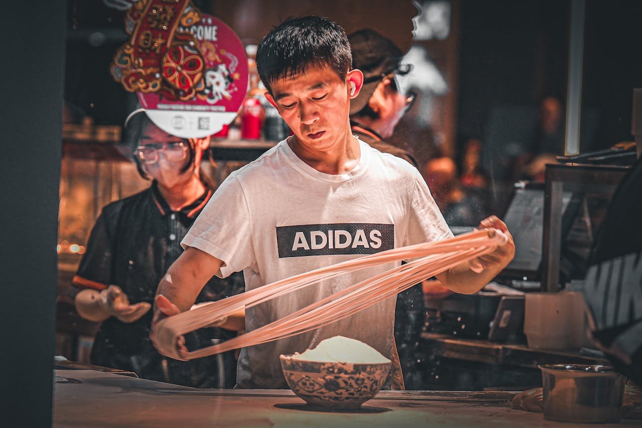 Chef expertly stretches noodles in a lively indoor setting, showcasing culinary precision.