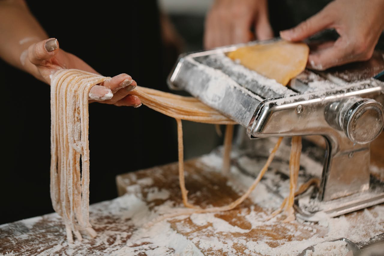 heros-img Crop anonymous female producing long noodle with metal pasta cutter on wooden table covered with flour